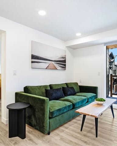 Modern living room with green velvet sofa, wood coffee table, and glass doors opening to a deck with mountain views