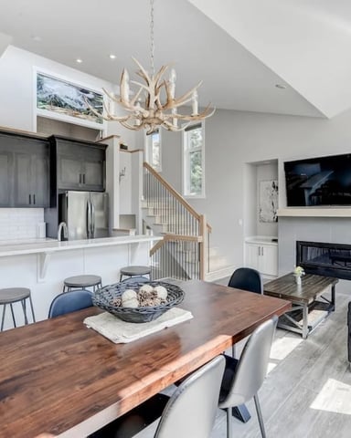 Open-plan kitchen and living room with white cabinetry, wood dining table, antler chandelier, fireplace, and staircase