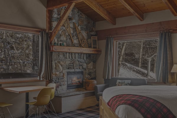 Rustic cabin bedroom with stone fireplace, exposed wood beams, large windows overlooking snowy forest, and plaid bedding.