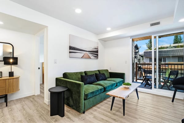 Modern living room with green velvet sofa, wood coffee table, and glass doors opening to a deck with mountain views