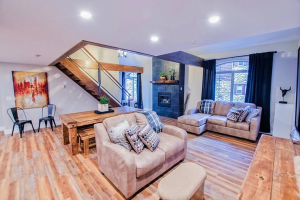 Open-plan living room with wooden staircase, beige seating, blue fireplace, and large windows with dark curtains