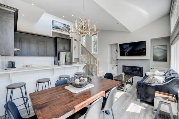 Open-plan kitchen and living room with white cabinetry, wood dining table, antler chandelier, fireplace, and staircase.