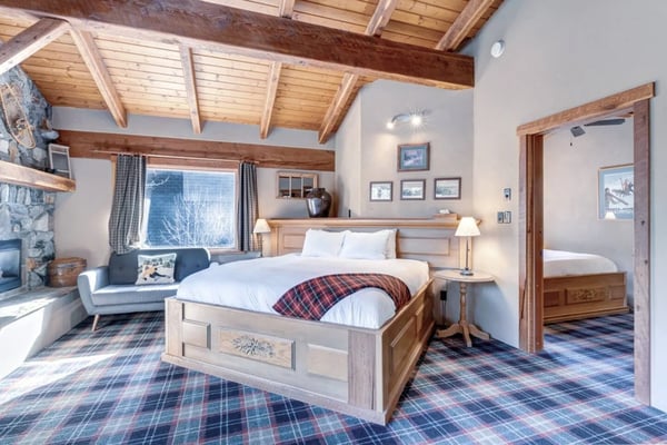 Spacious bedroom with wooden bed, exposed beam ceiling, stone fireplace, window with mountain view, and sitting area
