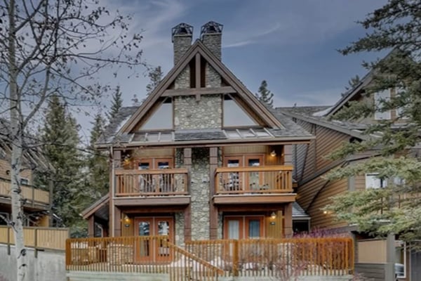 Modern mountain home with stone chimney, peaked roof, wooden decks, and surrounding evergreen trees